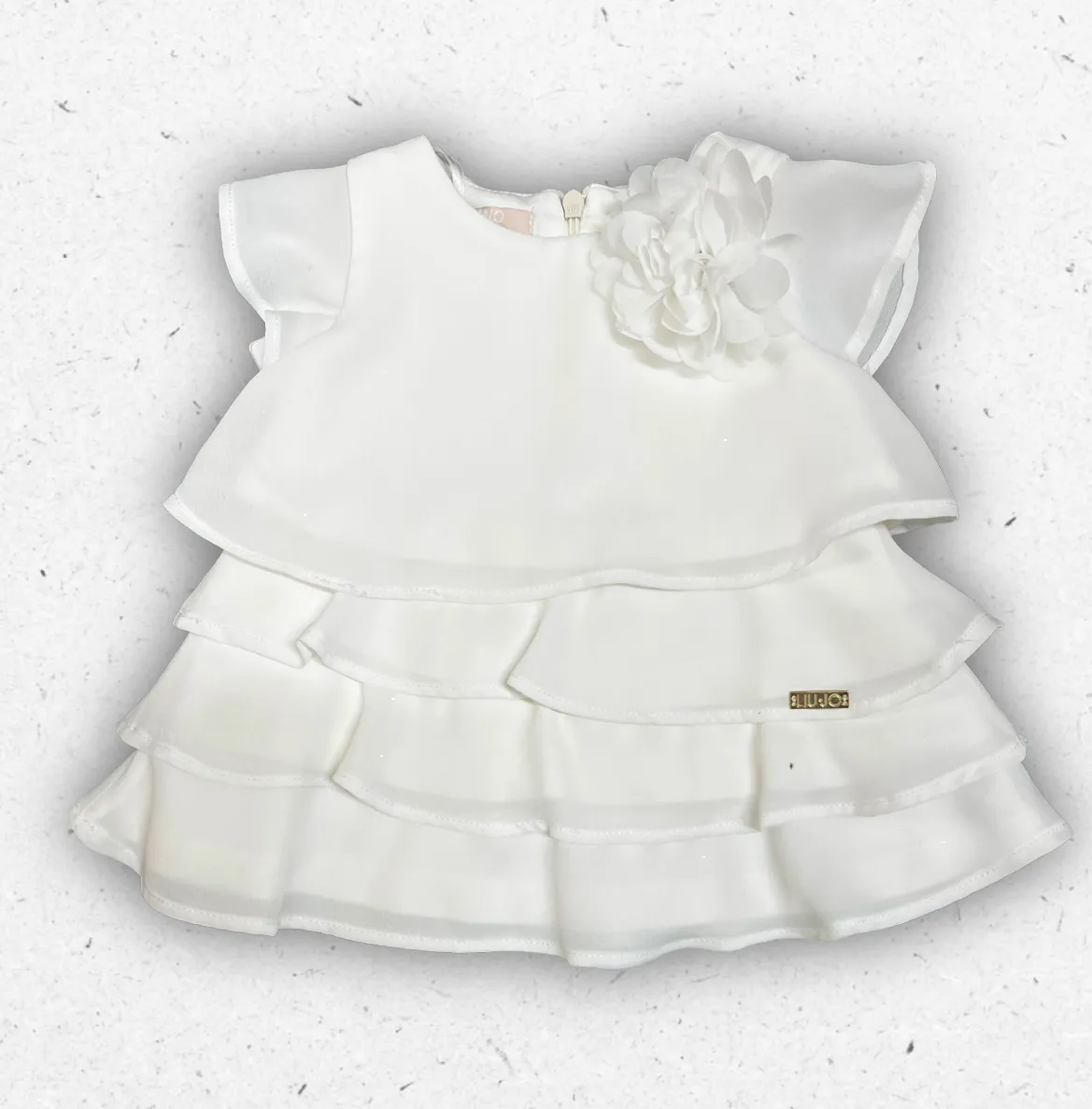 Abito bianco da cerimonia per neonata Liu Jo con design a balze sovrapposte, maxi fiore applicato sulla spalla e placchetta logo dorata.